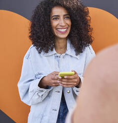 Lächelnde Frau, die ein Mobiltelefon hält und mit einer Freundin spricht, während sie an der Wand steht - SUF00653