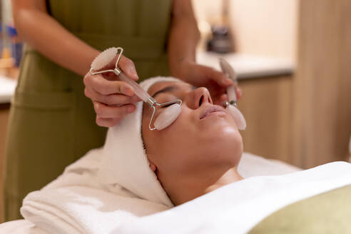 Kosmetikerin hält Edelstein-Roller auf das Gesicht einer jungen Kundin während einer Behandlung im Wellness-Center - OCMF01890