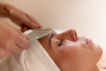 Beautician holding gua sha on female customer's forehead at health spa - OCMF01899