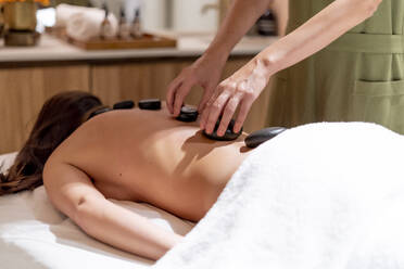 Massage therapist positioning stones on female customer's back at beauty spa - OCMF01920