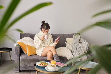 Smiling woman using smart phone while having coffee on sofa at home during winter - AODF00086