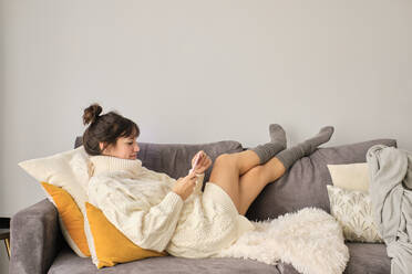 Woman using smart phone while relaxing on sofa at home during winter morning - AODF00087