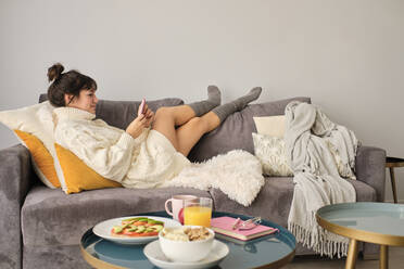 Woman using mobile phone while relaxing at home on winter morning - AODF00088