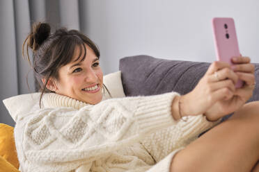 Lächelnde schöne Frau im Pullover nimmt Selfie durch Smartphone beim Entspannen zu Hause - AODF00091
