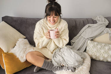 Glückliche Frau mit Kaffee im Becher, während sie im Winter zu Hause auf dem Sofa sitzt - AODF00092