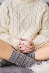 Frau im Pullover mit Kaffeetasse auf dem Sofa zu Hause sitzend - AODF00094