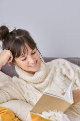 Lächelnde Frau liest ein Buch und entspannt sich auf dem Sofa zu Hause - AODF00103