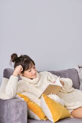 Woman reading book while reclining on sofa at home during winter - AODF00104