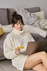 Smiling woman shopping online with credit card through laptop while sitting in living room at home - AODF00106