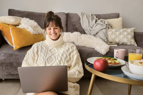 Smiling woman in sweater using laptop while sitting in living room at home - AODF00109