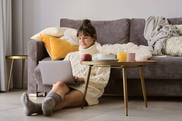 Frau im Pullover, die einen Laptop benutzt, während sie zu Hause im Wohnzimmer sitzt - AODF00111