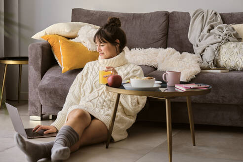 Woman in sweater having juice while using laptop at home - AODF00113