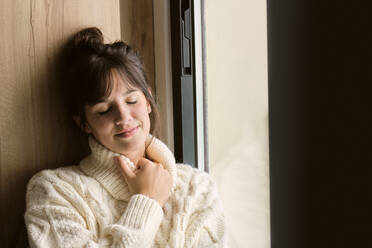 Nachdenkliche Frau mit geschlossenen Augen, die im Winter zu Hause am Fenster sitzt - AODF00117