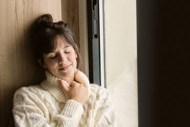 Thoughtful woman with eyes closed sitting by window at home during winter - AODF00117