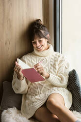 Lächelnde Frau schreibt in ein Buch, während sie zu Hause am Fenster sitzt - AODF00119