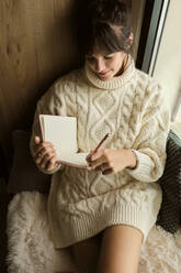 Smiling woman wearing sweater writing in book while sitting by window at home - AODF00120