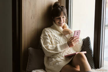 Woman in sweater smiling while video calling through mobile phone by window at home - AODF00126