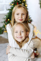 Lächelnde Schwestern umarmen sich mit einem Weihnachtsbaum im Hintergrund zu Hause - EYAF01447