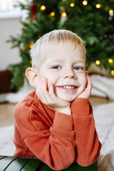 Close-up portrait of cute boy with hands on chin sitting with Christmas tree in background - EYAF01451