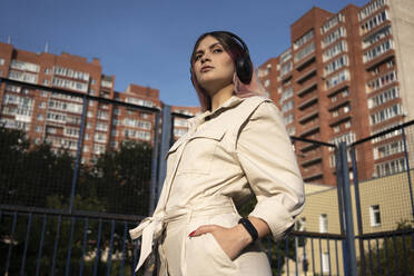 Hipster-Frau mit Händen in den Taschen hört Musik über Kopfhörer an einem sonnigen Tag - VPIF03328