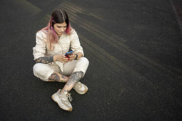 Modische Frau mit rosa Haaren, die ein Mobiltelefon benutzt, während sie auf einem Sportplatz sitzt - VPIF03340