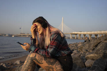 Smiling young woman with smart phone listening music on rock at beach during sunny day - VPIF03374