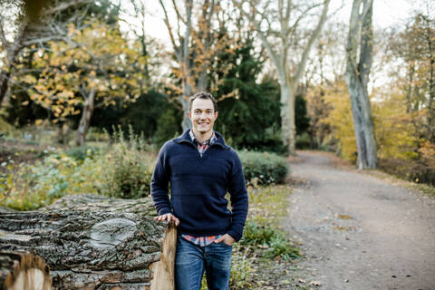 Lächelnder gut aussehender Mann mit Hand in der Tasche vor Bäumen in einem öffentlichen Park stehend, lizenzfreies Stockfoto