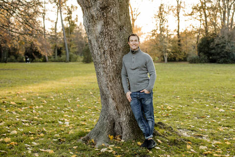 Smiling mid adult man standing by tree trunk in public park stock photo