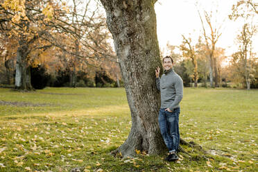 Lächelnder Mann an einem Baumstamm in einem öffentlichen Park im Herbst - KVF00150