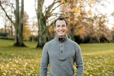 Cheerful handsome man standing in front of trees in public park during autumn - KVF00151