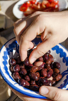 Hände einer Person, die schwarze Oliven aus einer Schale isst - EGBF00554