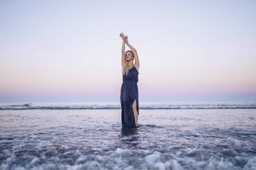 Modische Frau mit erhobener Hand am Strand Platja de Llevant bei Sonnenuntergang - RSGF00468