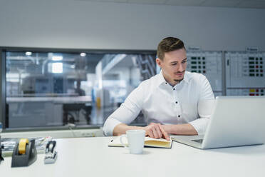 Businessman with diary working on laptop in factory office - DIGF13558