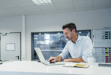 Männlicher Fachmann, der an einem Laptop arbeitet, während er im Sitzungssaal eines Fabrikbüros sitzt - DIGF13559