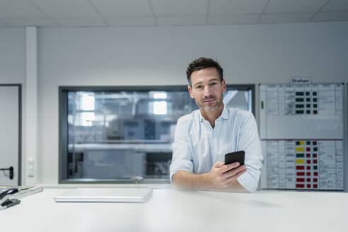 Männlicher Fachmann mit Mobiltelefon am Schreibtisch in einem Fabrikbüro - DIGF13561