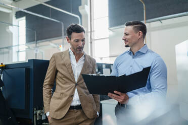 Smiling businessman with male coworker talking while holding file in industry - DIGF13588