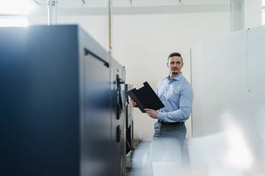 Male professional with file document looking away while standing by machine in factory - DIGF13590