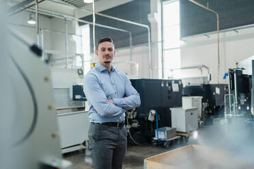 Confident businessman with arms crossed surrounded by machines in industry - DIGF13592