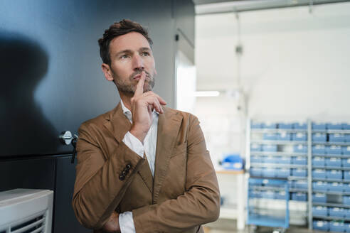 Nachdenklicher Geschäftsmann mit Finger auf den Lippen an einer Maschine in der Industrie - DIGF13596