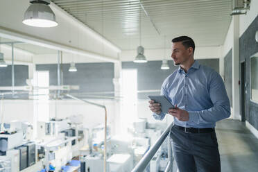 Mid adult businessman holding digital tablet by railing while looking around in industry - DIGF13606