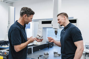 Smiling technician with male coworker discussing over machine part in industry - DIGF13615