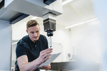 Männlicher Techniker mit Papier, der einen Bohrer in einer Maschine in einem Fabrikbüro befestigt - DIGF13619