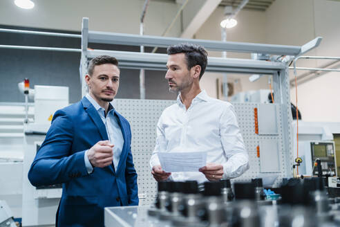 Geschäftsmann sieht einen männlichen Mitarbeiter an, während er über eine Maschine in einer Fabrik diskutiert - DIGF13641