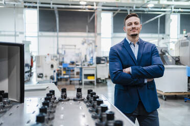 Smiling businessman with arms crossed by manufacturing equipment in factory - DIGF13645