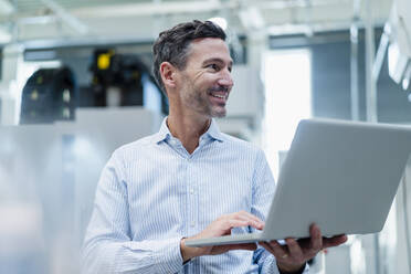 Lächelnder männlicher Fachmann mit Laptop, der in einer Fabrik wegschaut - DIGF13682