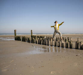 Woman with arms outstretched standing on wooden posts at beach against clear sky during vacation - UUF22328