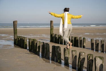 Mature woman with arms outstretched balancing on wooden posts at beach - UUF22329