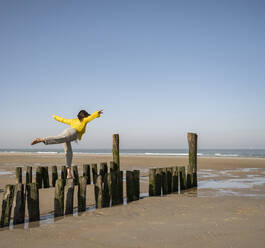 Mature woman with arms outstretched balancing on wooden posts at beach against clear sky - UUF22330