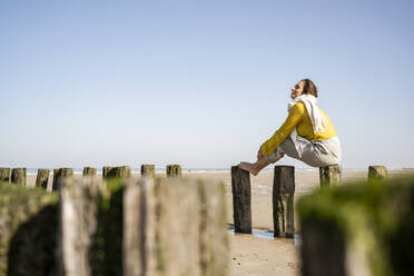 Mature woman sitting on wooden post at beach against clear sky - UUF22331