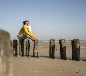 Frau sitzt auf Holzpfosten am Strand gegen den klaren Himmel an einem sonnigen Tag - UUF22334